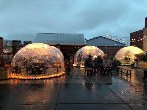 Three illuminated geodesic domes on a rainy evening, with people gathered around a fire pit and string lights overhead.