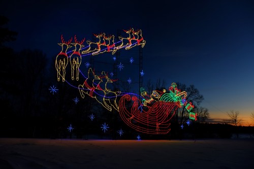 Colorful holiday lights depict Santa in a sleigh pulled by reindeer against a twilight sky. Snow-covered ground below.