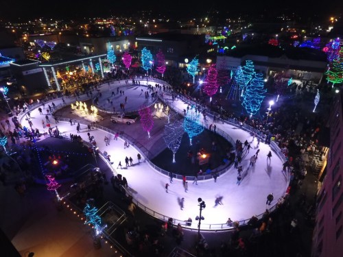 Aerial view of a bustling ice skating rink at night, surrounded by colorful holiday lights and a large crowd.