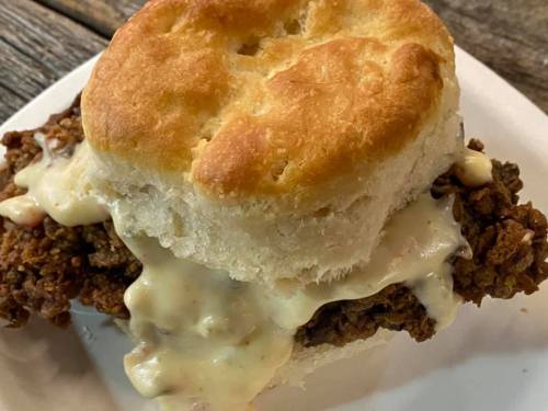 A close-up of a biscuit sandwich filled with fried chicken and creamy gravy on a white plate.