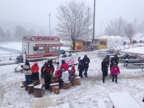 Snowy scene with people in winter clothing gathered around logs, food trucks in the background, and snow falling.