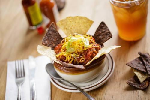 A bowl of chili topped with cheese and green onions, served with tortilla chips and a glass of iced tea.
