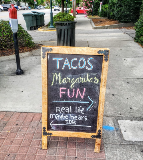 A chalkboard sign on the sidewalk advertising tacos, margaritas, and fun, with playful notes about bears and uncertainty.