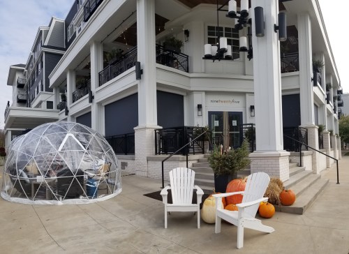 A cozy outdoor dining area with a geodesic dome, pumpkins, and chairs in front of a modern building.