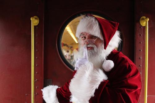 Santa Claus in a red suit and hat, holding a finger to his lips, standing by a round window.