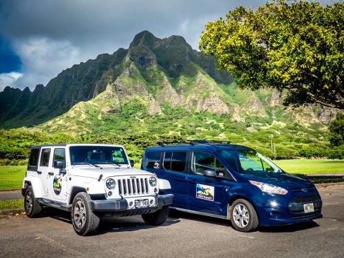 Two parked vehicles, a white Jeep and a blue van, with lush green mountains in the background under a partly cloudy sky.