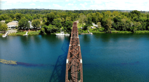 Aerial view of a rusty bridge spanning a calm river, surrounded by lush greenery and boats along the shore.