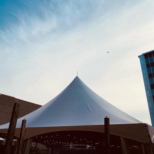 A large white tent with a pointed top against a blue sky, surrounded by buildings and string lights.
