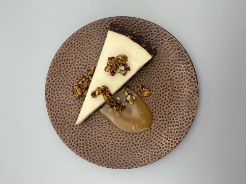 A slice of cheesecake topped with nuts on a textured brown plate, accompanied by a drizzle of caramel sauce.