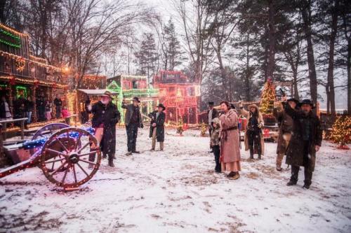 A snowy scene with people in winter attire, festive lights, and decorated buildings, celebrating in a holiday setting.