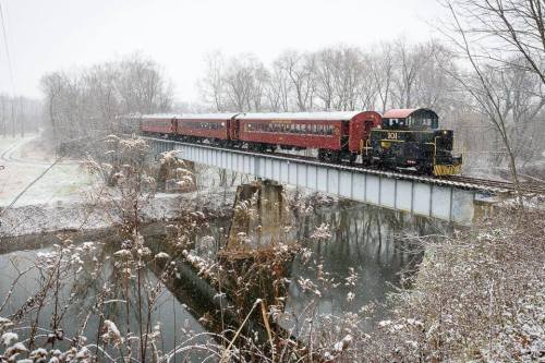 A vintage train crosses a snowy bridge over a river, surrounded by bare trees and a winter landscape.
