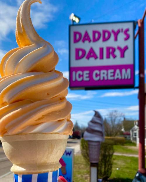 A soft-serve ice cream cone in front of a colorful sign for Daddy's Dairy Ice Cream.