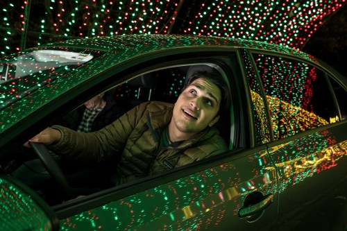 A man smiles inside a car surrounded by colorful holiday lights.