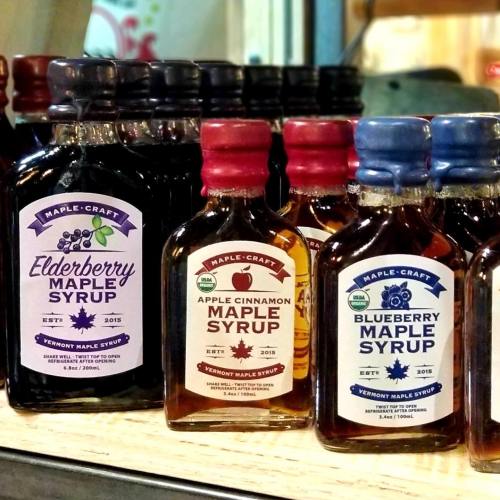 Bottles of flavored maple syrup, including elderberry, apple cinnamon, and blueberry, displayed on a shelf.