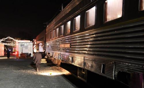 A person walks beside a lit train car at night, with festive lights in the background.
