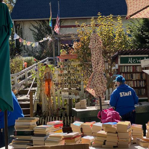 A vibrant outdoor book sale with stacks of books, colorful flags, and a sign for various attractions in a garden setting.