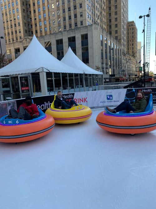 Three people sit in colorful bumper cars on an ice rink, with a tent and city buildings in the background.