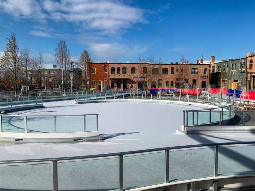 An empty outdoor ice skating rink surrounded by buildings and trees under a clear blue sky.