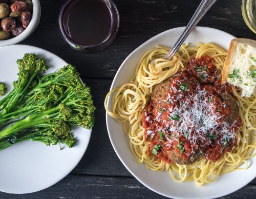 A plate of spaghetti with meatballs and marinara sauce, garnished with cheese, alongside broccolini and a glass of red wine.