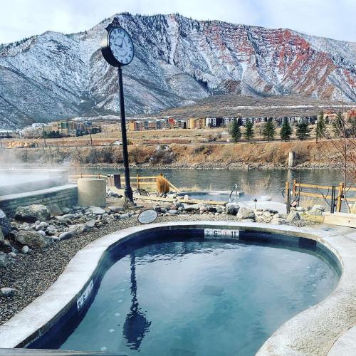 A scenic view of a hot spring with a clock, mountains in the background, and snow-covered peaks.