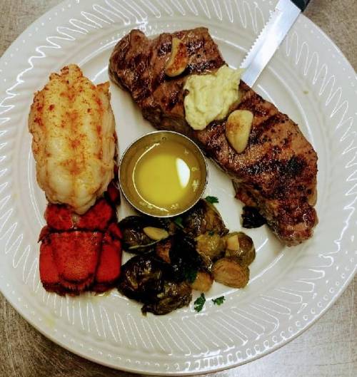 A plate featuring grilled steak with garlic butter, lobster tail, Brussels sprouts, and a small bowl of olive oil.