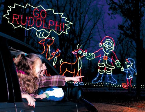 A girl leans out of a car, excitedly pointing at colorful holiday lights depicting Santa, Rudolph, and an elf.