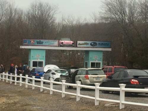 A drive-thru with a pink car on the roof, surrounded by parked vehicles and a white fence in a wooded area.