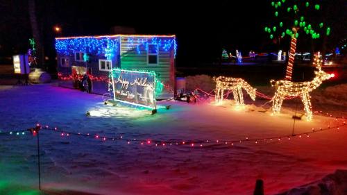 A festive scene with a decorated trailer and illuminated reindeer in a snowy landscape, surrounded by colorful lights.