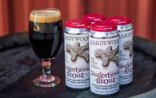 A glass of dark stout next to a four-pack of Hardywood Gingerbread Stout cans on a wooden barrel.