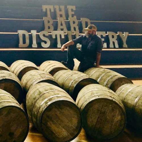 A man sits on whiskey barrels in front of a sign that reads "The Bard Distillery."