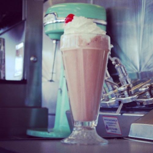 A tall glass of chocolate milkshake topped with whipped cream and a cherry, with a retro mixer in the background.