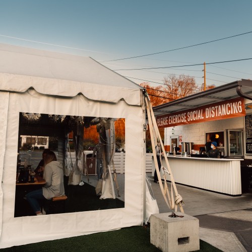 A tented outdoor dining area with a sign urging social distancing, featuring a bar and patrons inside.