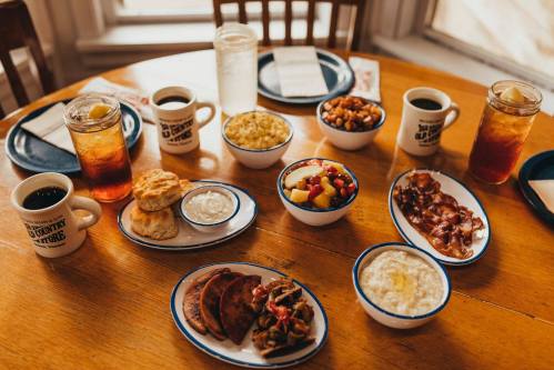 A table set with various breakfast dishes, including biscuits, bacon, fruit, and drinks in mugs and glasses.