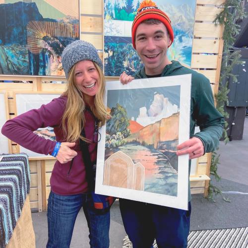 A man and woman smile while holding a framed artwork at a market, surrounded by colorful art displays.