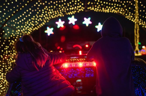 Two children lean on a car, watching colorful holiday lights and stars in a festive nighttime display.