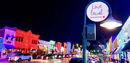 Colorful holiday lights adorn buildings along a street, with a sign reading "Love local Rochester" visible.