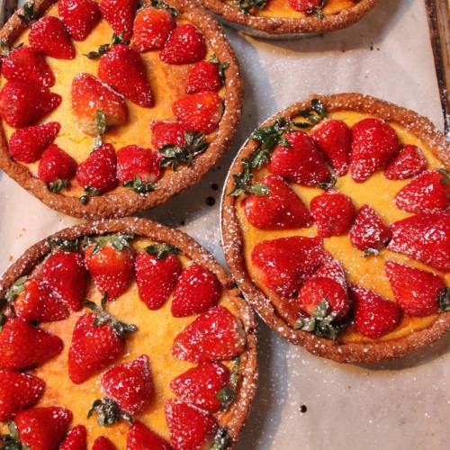 Four strawberry tarts with a creamy filling, topped with fresh strawberries and dusted with powdered sugar.