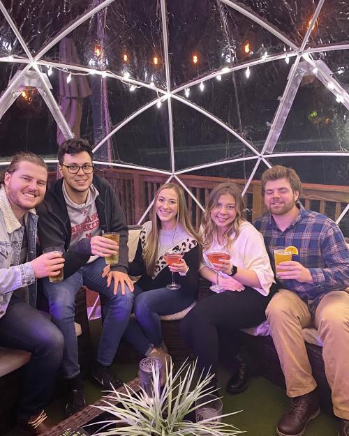 A group of five friends sitting together in a cozy, illuminated dome, each holding a cocktail and smiling.