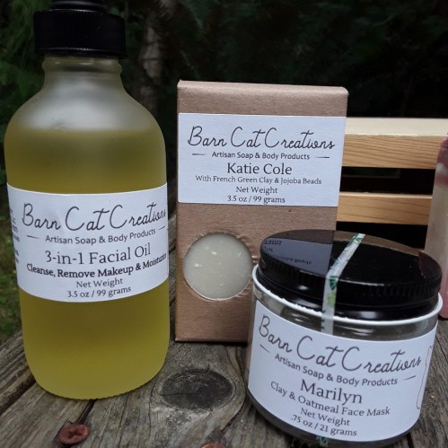 A bottle of facial oil and two soap bars labeled "Barn Cat Creations" on a wooden surface surrounded by greenery.