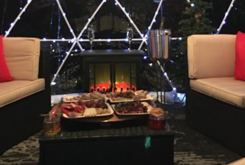 Cozy outdoor seating under a lit dome, featuring a small fireplace and a table with assorted food and drinks.