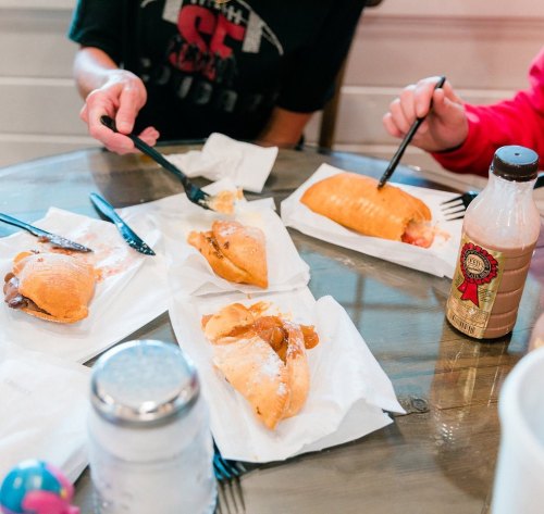 A table with several fried pastries on paper plates, forks in hand, and a bottle of drink nearby.
