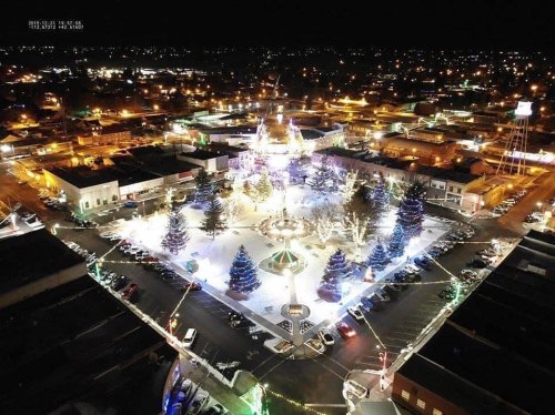 Aerial view of a festive town square illuminated with holiday lights and decorated trees at night.