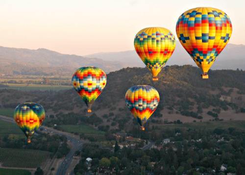 Colorful hot air balloons float over a scenic landscape with mountains and vineyards during sunset.