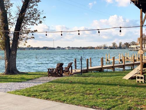 A serene lakeside view with wooden chairs, a dock, and string lights under a blue sky with fluffy clouds.