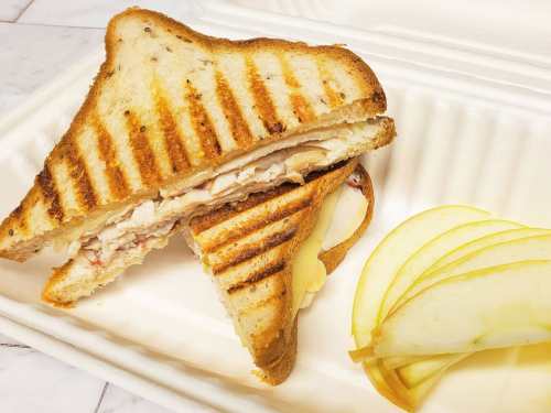 A grilled sandwich with sliced turkey and cheese, served with apple slices on a biodegradable container.
