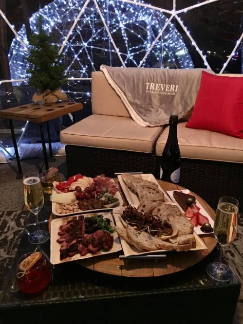 A cozy dining setup featuring a charcuterie board, drinks, and a festive backdrop of lights and a plush sofa.