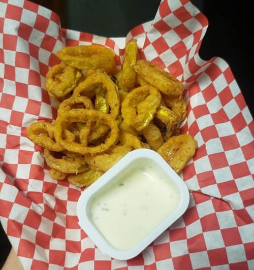 A serving of crispy fried snacks, including onion rings and plantains, served with a small cup of dipping sauce.