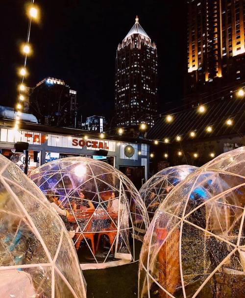 A cozy outdoor dining area with transparent igloos, string lights, and a city skyline at night.