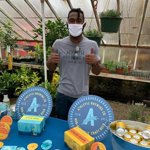 A person wearing a mask gives a thumbs up at a brewery booth with craft beer products and plants in the background.