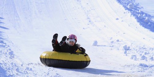 A child in a pink helmet slides down a snowy hill on a yellow and black snow tube, enjoying a winter day.